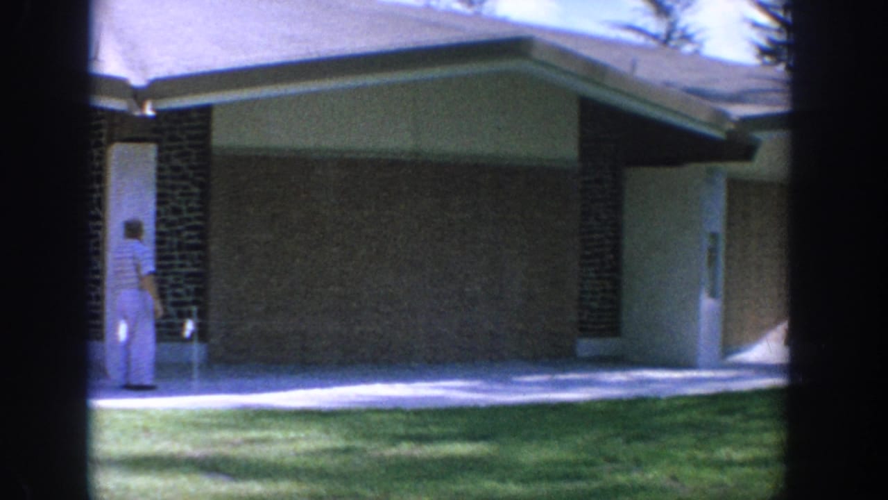 A man stands beside a doorway on a low building with a lawn in front.
