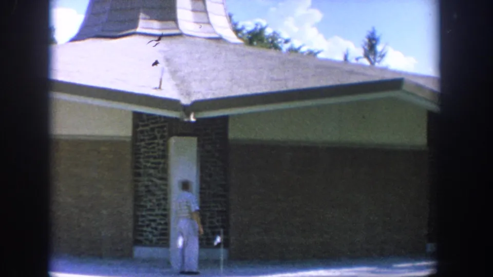 1964 Wisconsin 8mm frame of a man near a doorway under a low roofline, with brick columns and lawn visible.