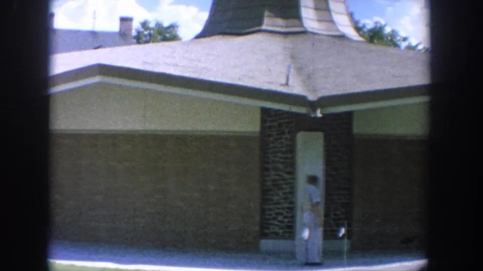 1964 Wisconsin home-movie frame showing a man at a building entrance with lawn, brickwork, and soft 8mm color.