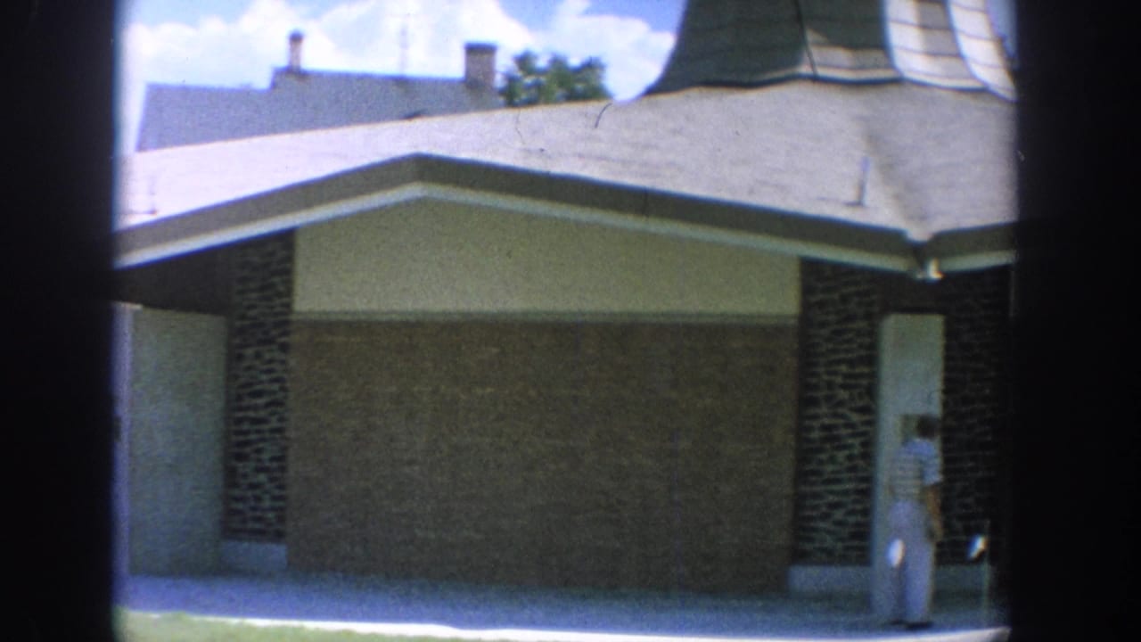 1964 Wisconsin 8mm frame showing a modern brick-and-stucco building exterior with a woman partly visible near a shaded entry.
