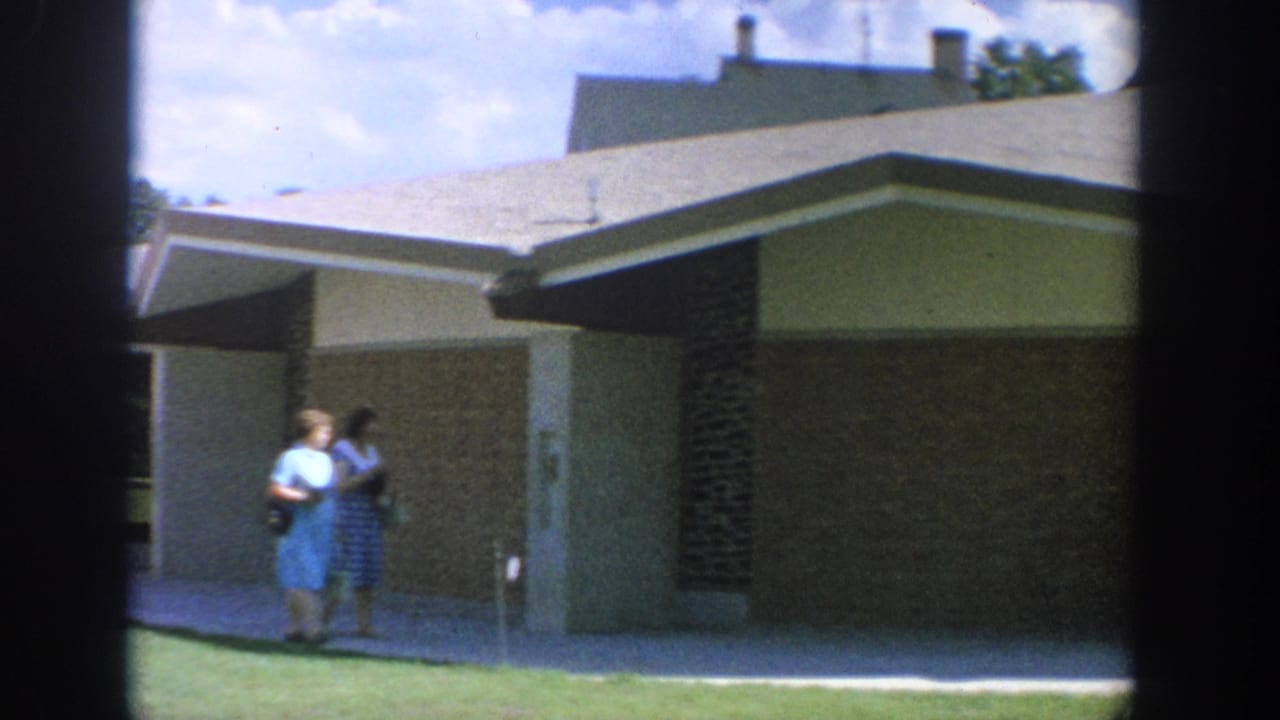 Two women walk together on grass beside a modern brick building on a sunny day.