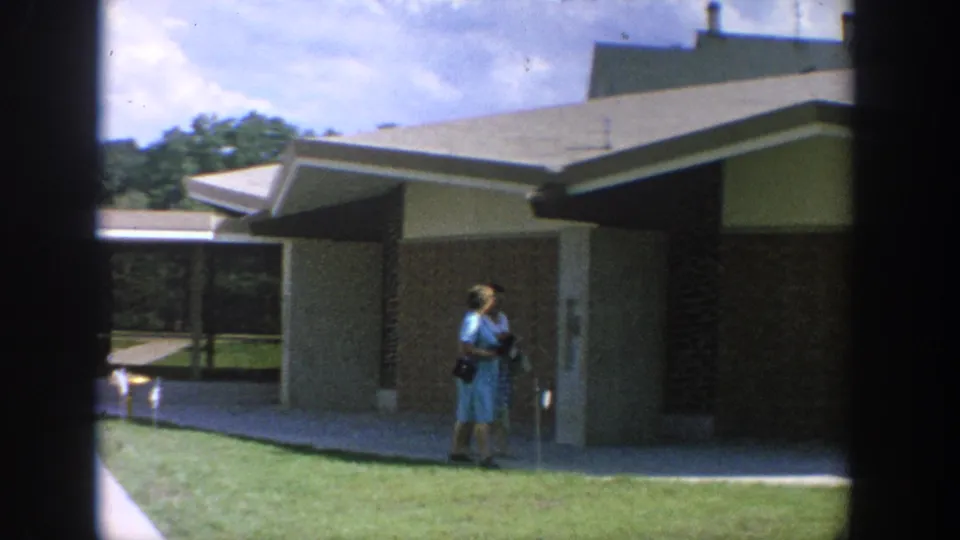 A woman in a light blue dress and dark purse pauses near the lawn outside a modern brick building in 1964 Wisconsin footage.