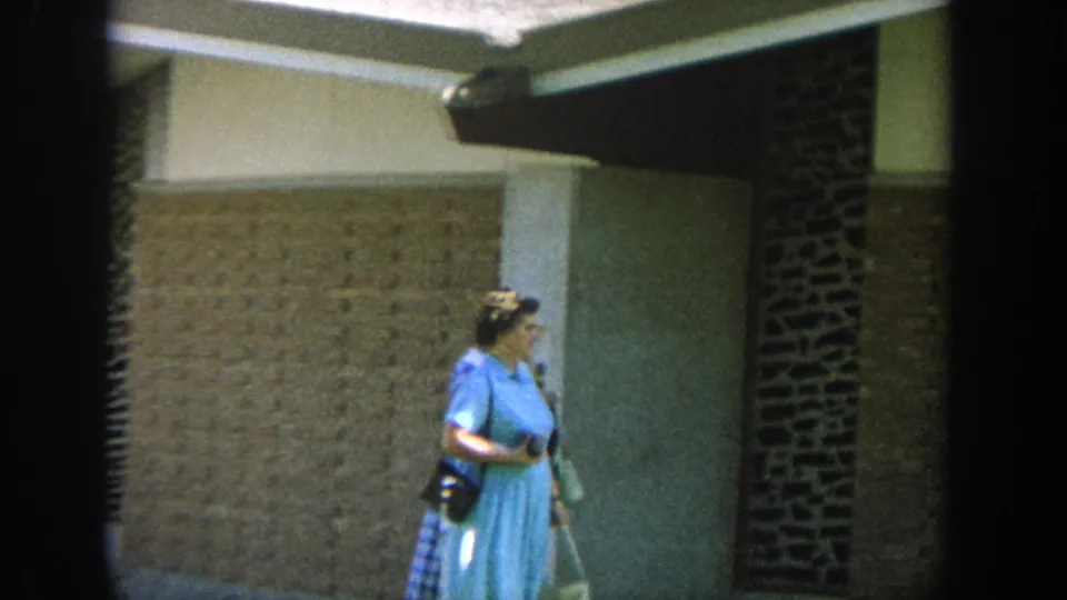 Closer 8mm frame of a woman in a light blue dress carrying a dark purse beside a brick-and-stucco wall in Wisconsin, 1964.