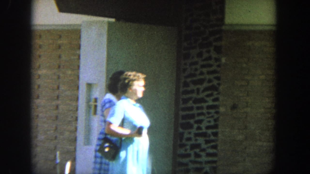1964 Wisconsin 8mm frame of an older woman in a light blue dress passing a brick building doorway in bright sun.