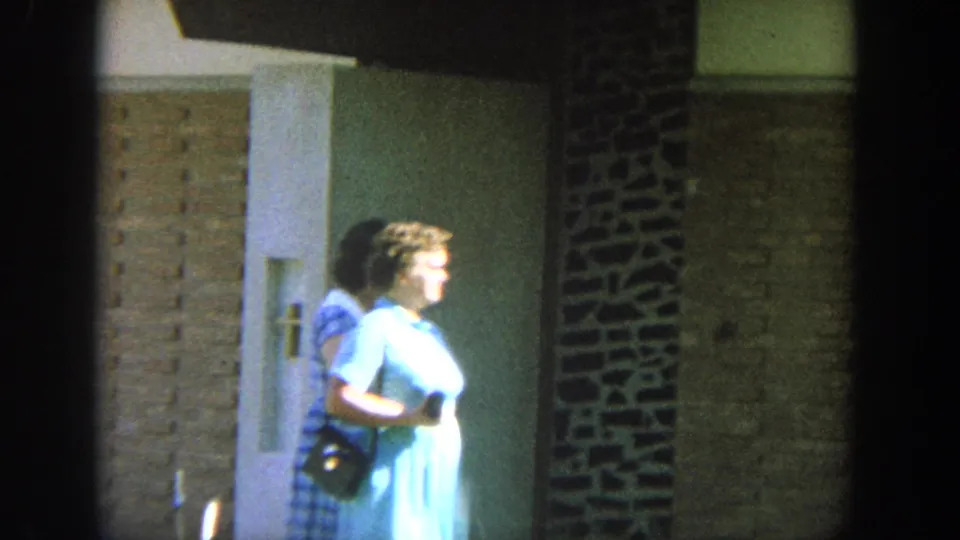 1964 Wisconsin 8mm frame of an older woman in a light blue dress passing a brick building doorway in bright sun.