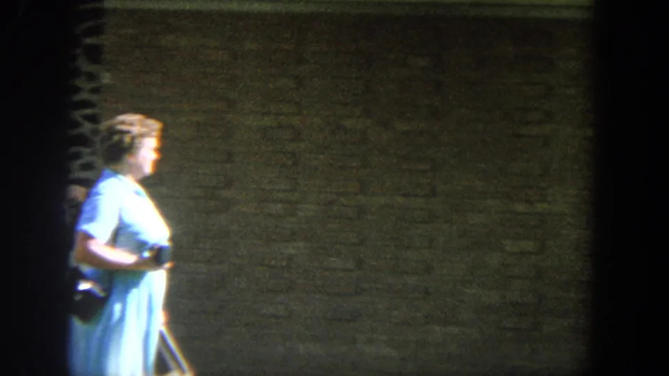 1964 Wisconsin home-movie frame showing an older woman in a blue dress moving past a brick exterior in strong sunlight.