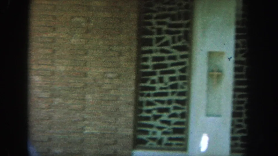 1964 Wisconsin 8mm frame with a brick exterior, doorway, and a partially visible woman in blue at the left edge.