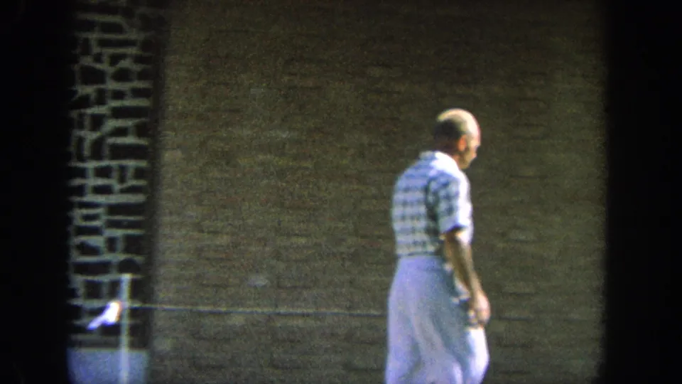 1964 Wisconsin home-movie frame of an older man in a plaid shirt walking along a brick building exterior.