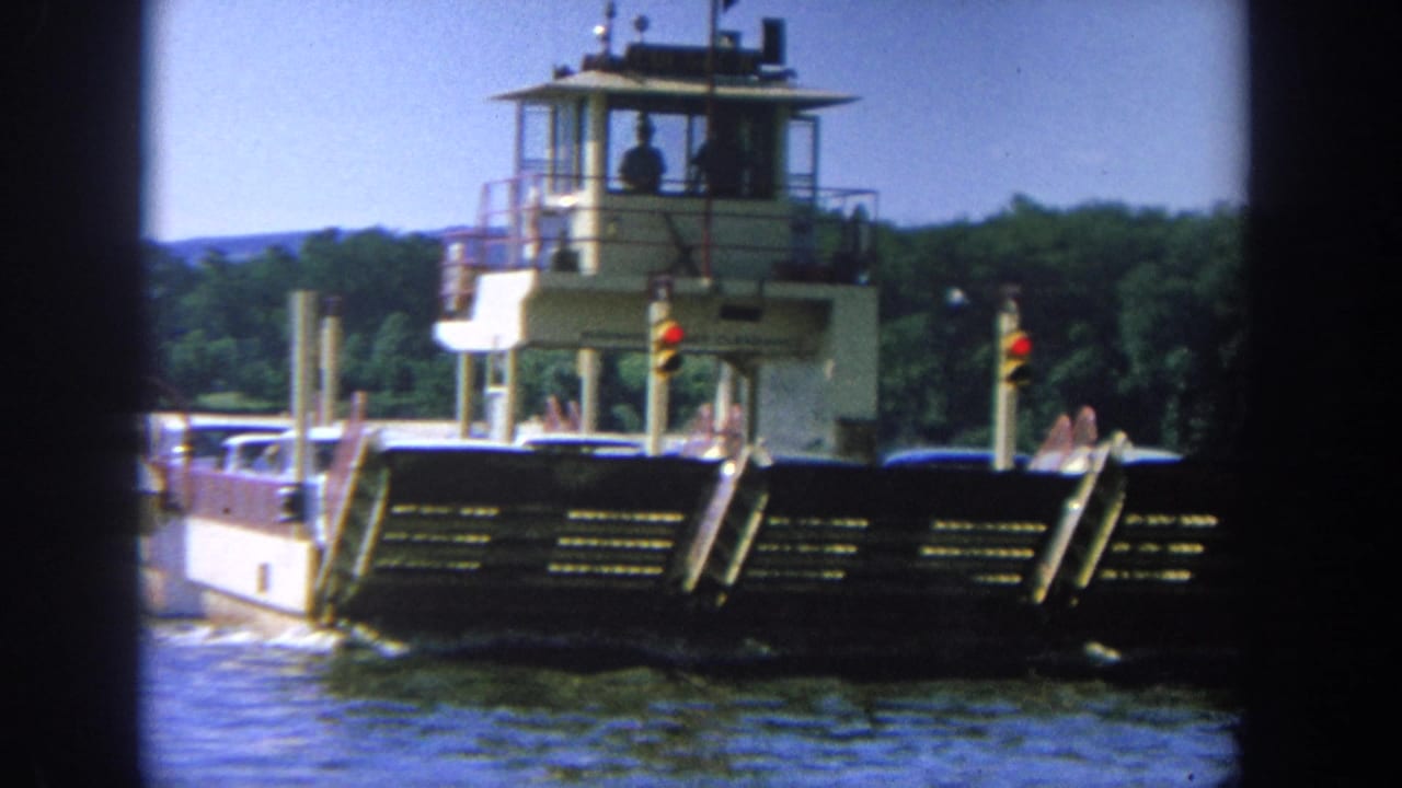 1964 Wisconsin home-movie frame showing a ferry-like barge with cars on deck and a pilot house above the river.