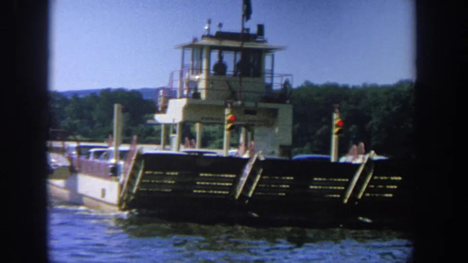 Side view of a 1964 Wisconsin river barge carrying cars, with a person silhouetted in the elevated control cabin.