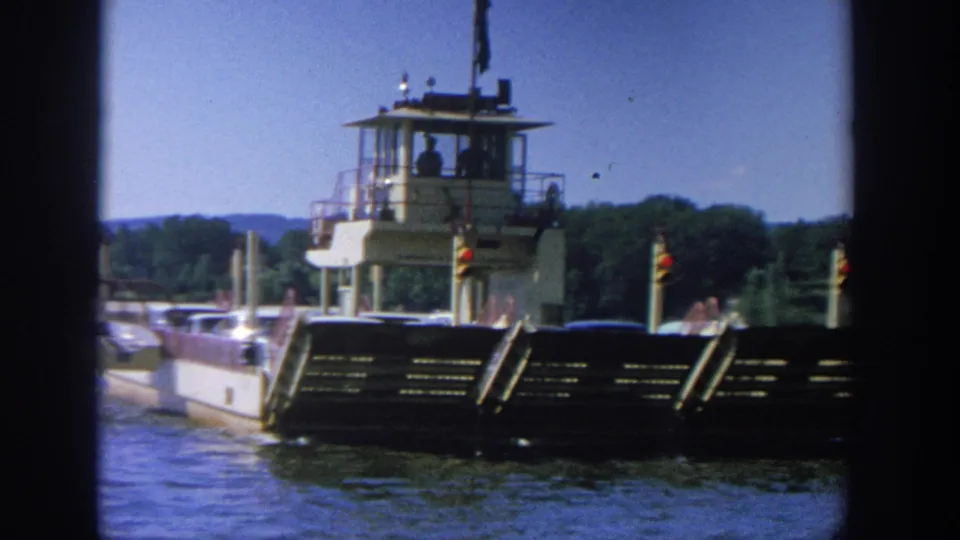 1964 Wisconsin home-movie still of a ferry-like barge on the river, with deck cars, passengers, and wooded shoreline behind.