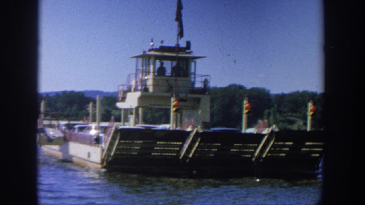 1964 Wisconsin 8mm frame of a river ferry carrying cars, with a white superstructure, dark ramp, and wooded shoreline behind it.