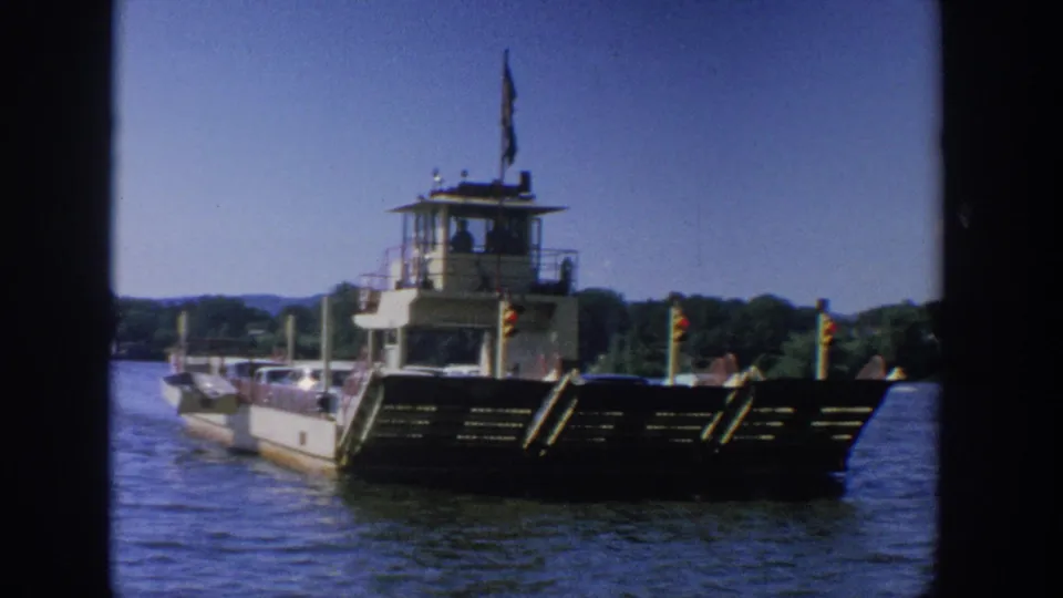 1964 Wisconsin 8mm frame of a ferry angled across blue water, showing its car deck, dark bow ramp, and raised wheelhouse.
