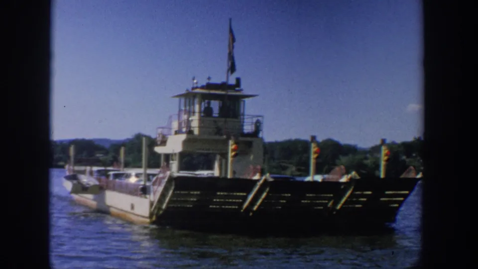 1964 Wisconsin home-movie frame showing the ferry front and control tower, with cars on deck and a wooded riverbank in the background.