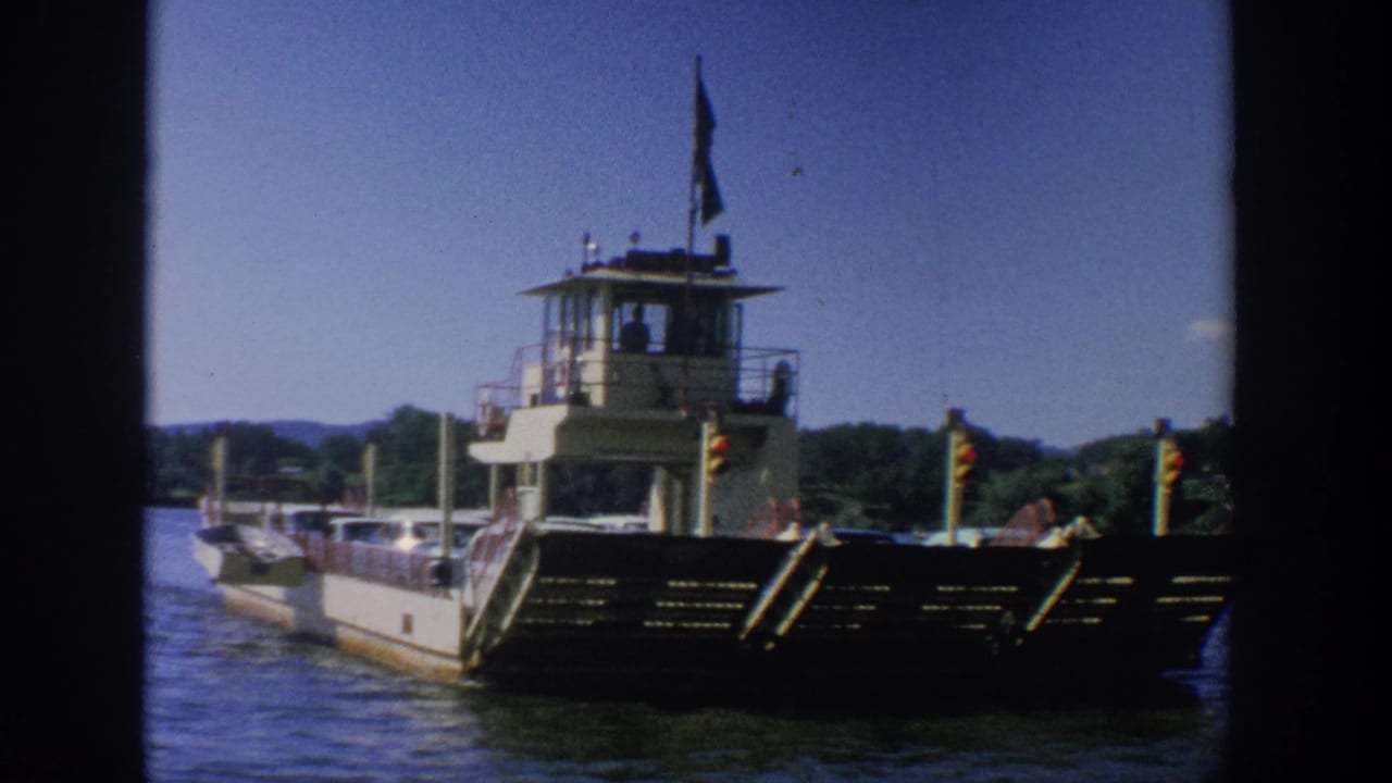 1964 Wisconsin 8mm view of a ferry boat on a river, with its bow ramp, pilothouse, flags, and shoreline visible.