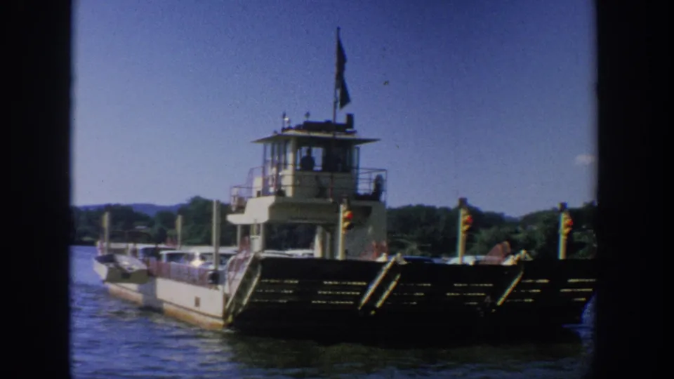 1964 Wisconsin 8mm view of a ferry boat on a river, with its bow ramp, pilothouse, flags, and shoreline visible.