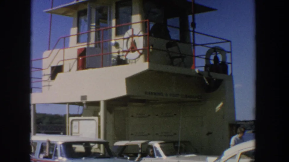 1964 Wisconsin ferry view showing cars on the lower deck, flags over the cabin, red railings, and a visible passenger or crew member.