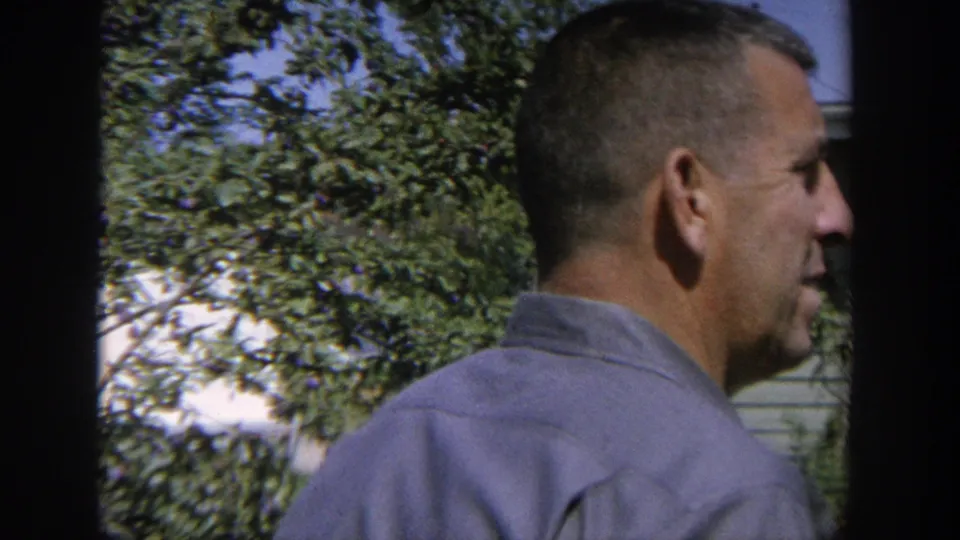 1964 Wisconsin home-movie frame showing an adult man in side profile near leafy trees with a house exterior behind him.