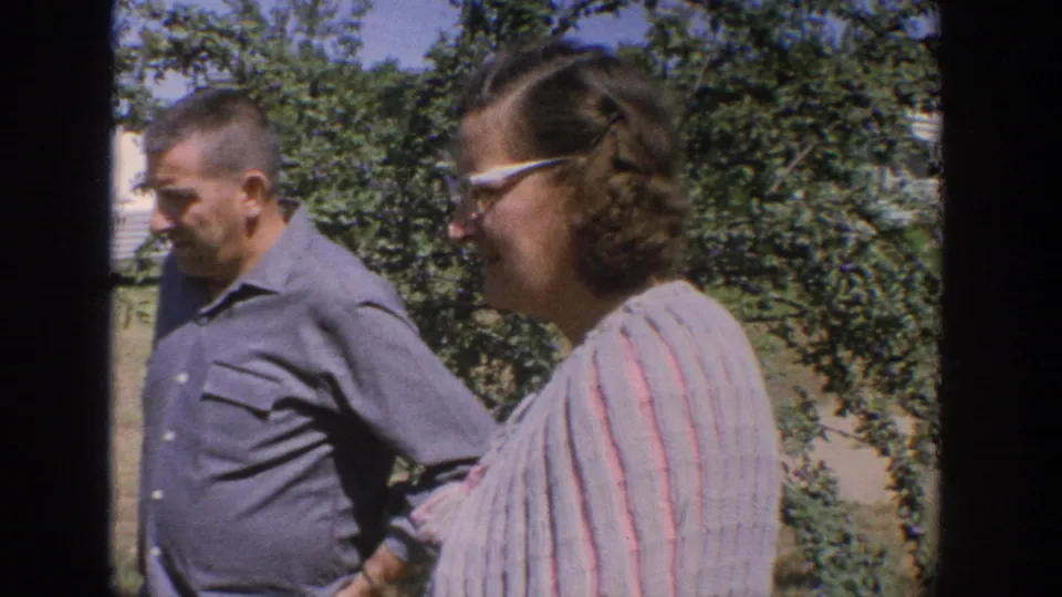 1964 Wisconsin home-movie frame showing a man and woman in a garden yard, with shrubs and a white trailer behind them.