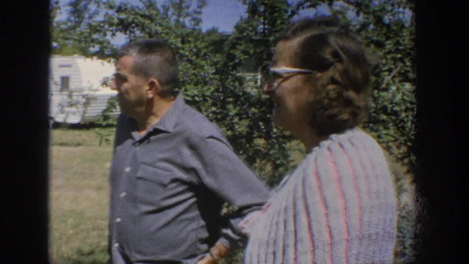 1964 Wisconsin 8mm frame of an adult couple standing in a yard near garden shrubs and a white trailer.