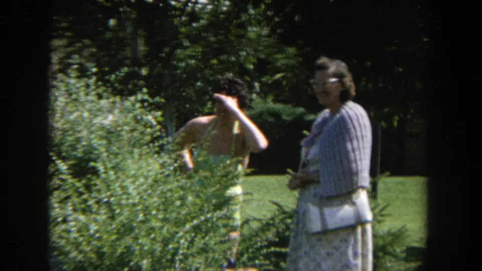 1964 Wisconsin 8mm frame with a woman in a yellow bathing suit behind a bush and another woman standing nearby in a dress and cardigan.