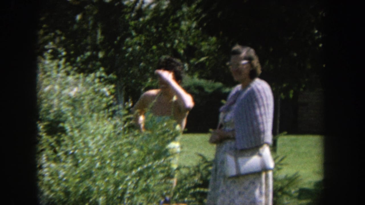 1964 Wisconsin home-movie frame showing one woman shielding her face near a bush while another woman watches in a sunlit garden.