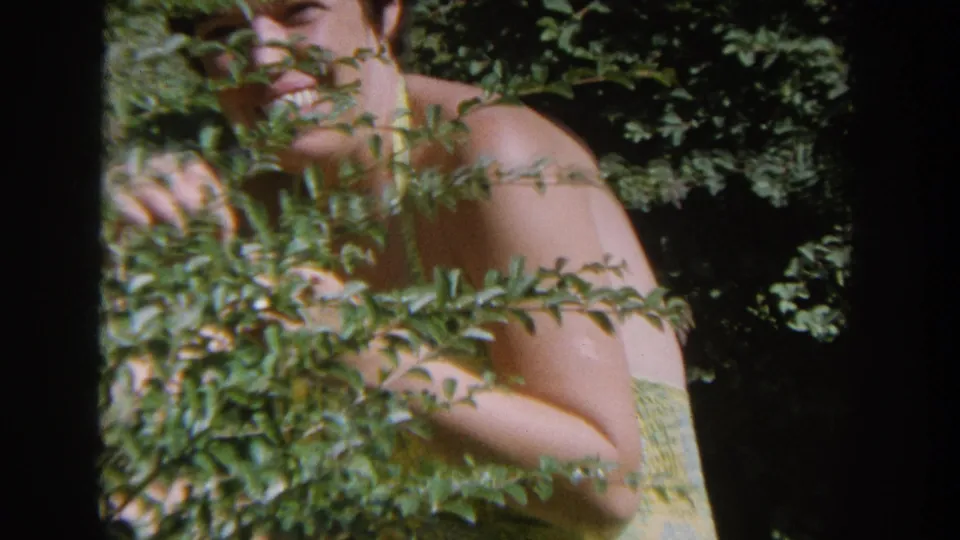 Close 1964 Wisconsin 8mm view of a woman in a yellow bathing suit partly obscured by dense green branches.