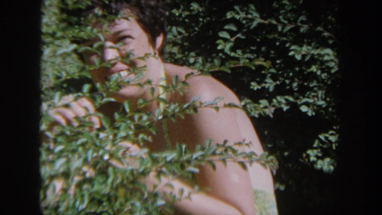 In 1964 Wisconsin home-movie footage, a woman in a yellow swimsuit peers through dense green shrubs in sunlight.