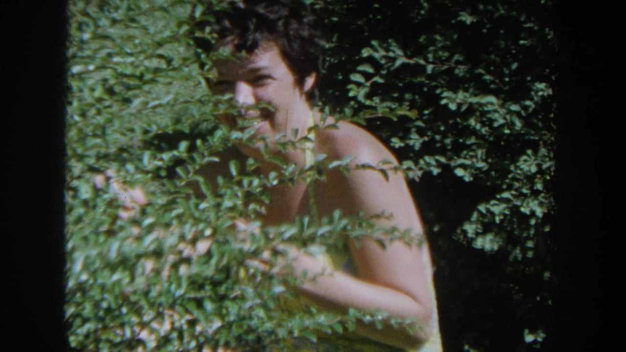Smiling woman in a yellow swimsuit stands behind a bush with dense greenery around her.