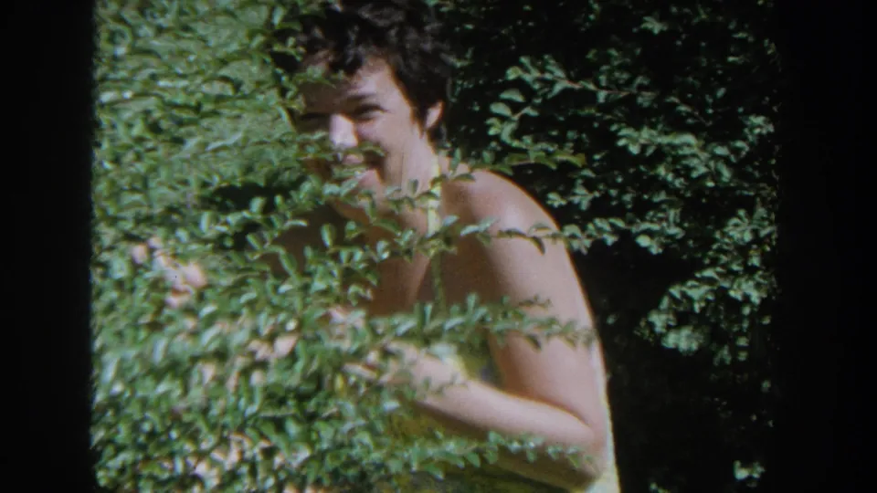 A smiling woman in a yellow swimsuit looks toward the camera from behind thick foliage in 1964 Wisconsin footage.