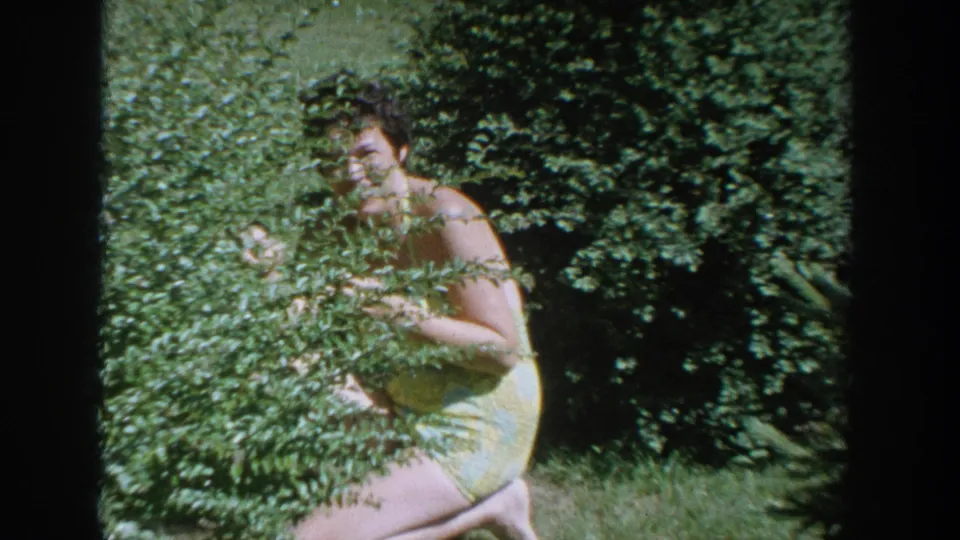 The woman remains partly concealed by a bush, framed by grass and thick garden greenery in the 1964 Wisconsin home movie.