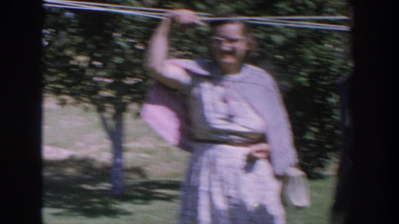 1964 Wisconsin home-movie frame showing a woman in a light dress and glasses standing in a grassy yard near a clothesline.