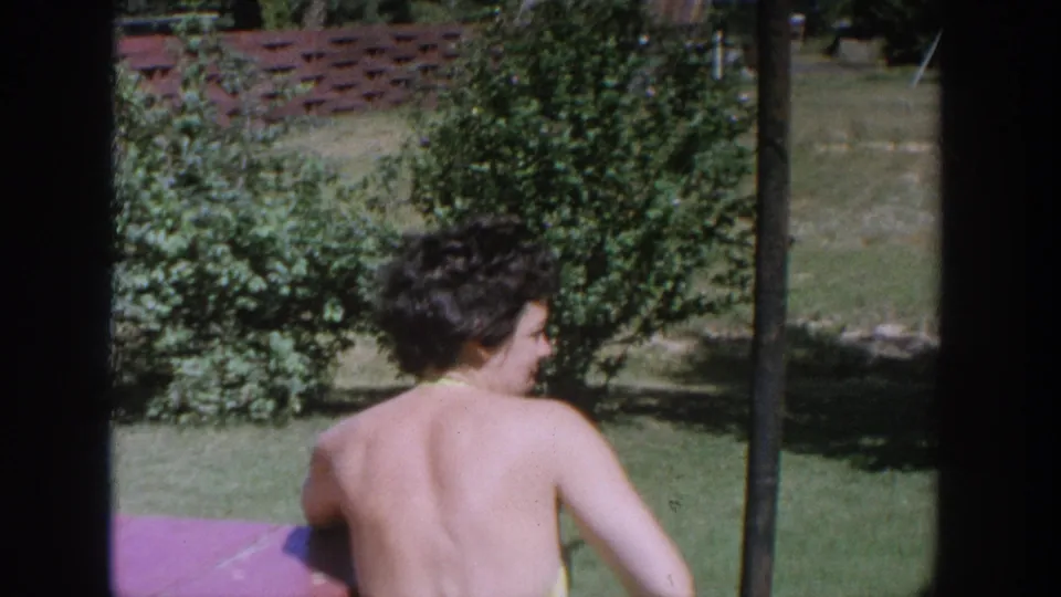 1964 Wisconsin home-movie frame showing a woman from behind near a red picnic table, with shrubs and lawn in the background.