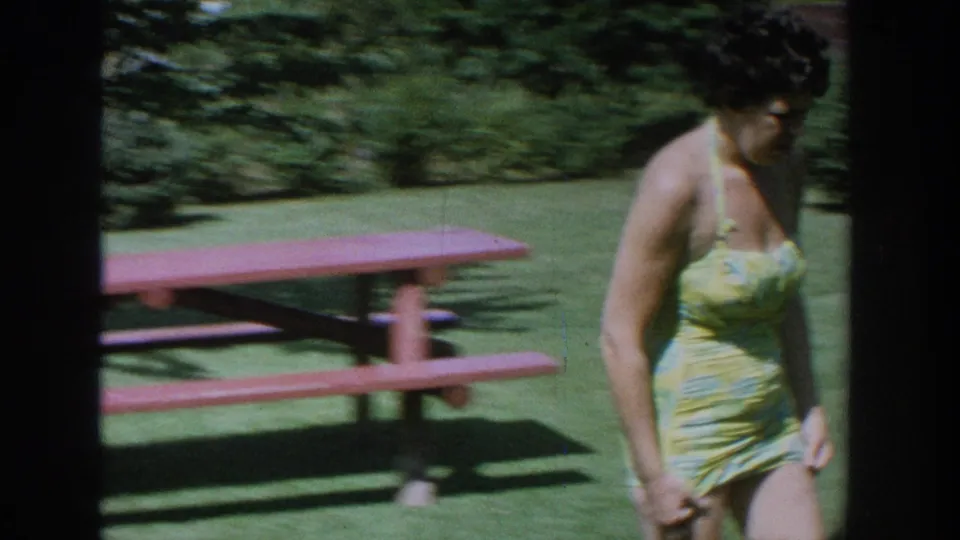 1964 Wisconsin home-movie frame showing a woman in a yellow-green swimsuit walking beside a red picnic table on a sunny lawn.