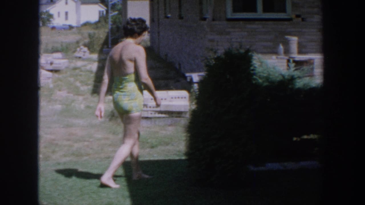 The woman walks barefoot away from the camera beside a brick house, with lawn, shrubbery, and yard objects visible.