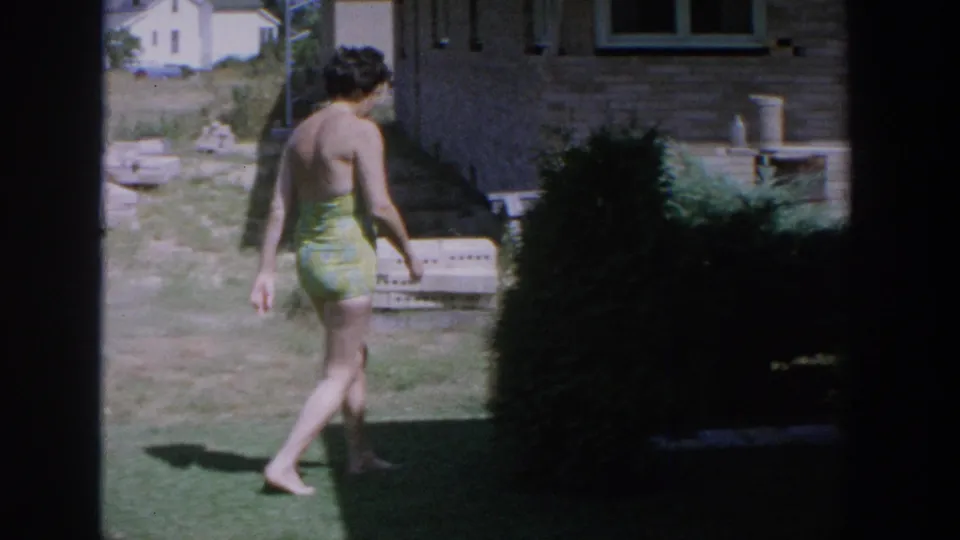 The woman walks barefoot away from the camera beside a brick house, with lawn, shrubbery, and yard objects visible.