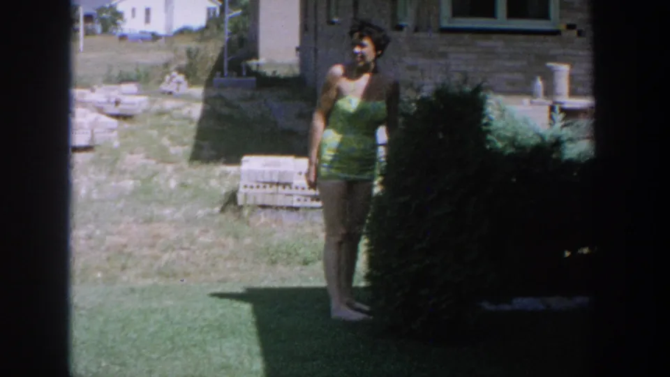 The woman pauses near a shrub in a Wisconsin backyard, facing partly toward the camera in a yellow-green swimsuit.