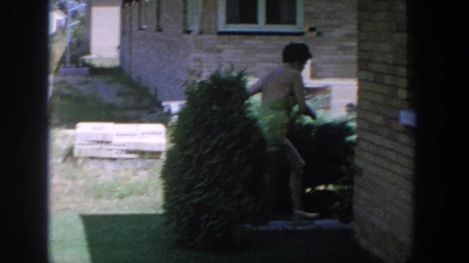 At the end of the clip, the woman moves into shade beside the brick house and shrub, partly obscured by the frame edge.