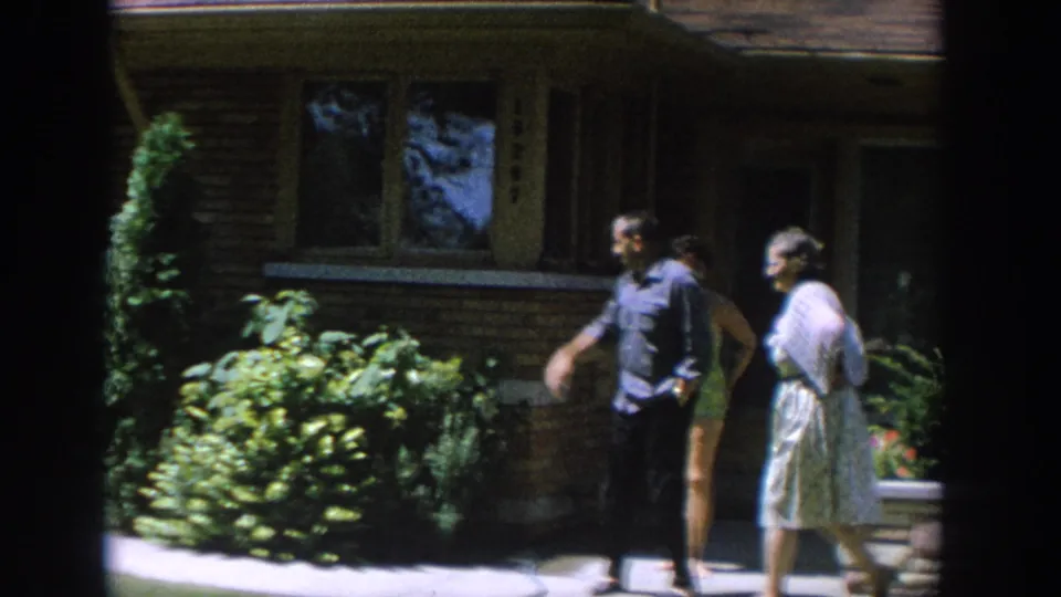 1964 Wisconsin home-movie frame showing a man and woman standing outside a brick house with shrubs and flowers nearby.