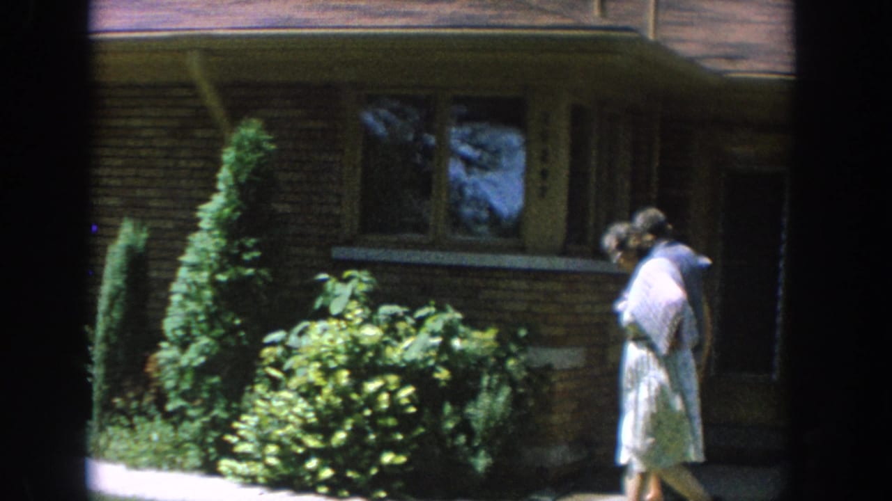 1964 Wisconsin home-movie frame showing a woman in a light dress walking past shrubs in front of a brick ranch-style house.
