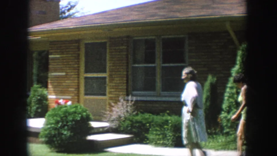1964 Wisconsin home-movie frame showing a woman in a light dress near the porch as another adult stands by the landscaping.