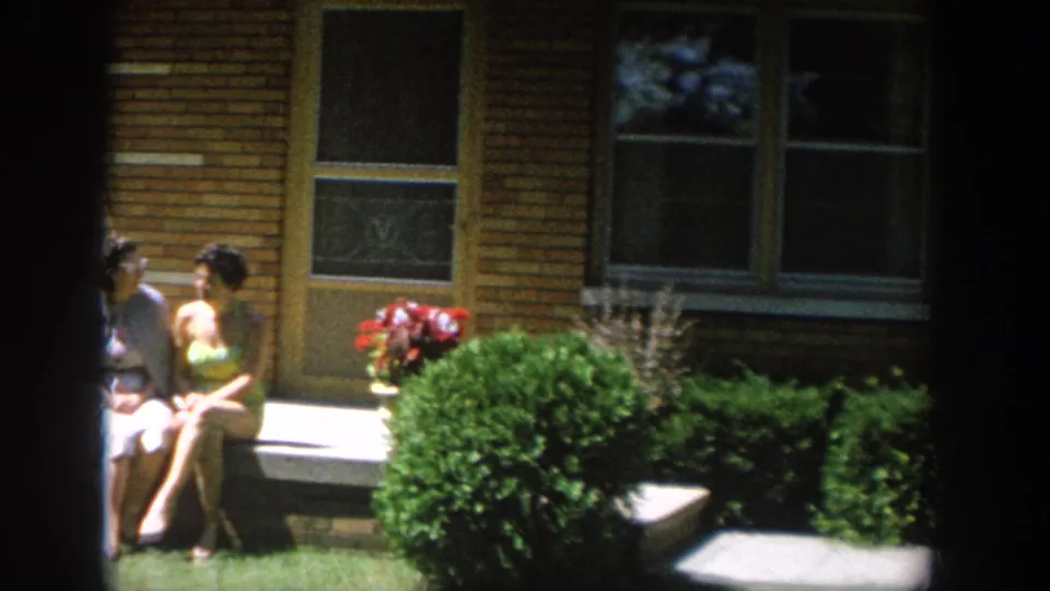 1964 Wisconsin frame showing women near a brick house porch, with one seated by the steps and flowers beside the entrance.
