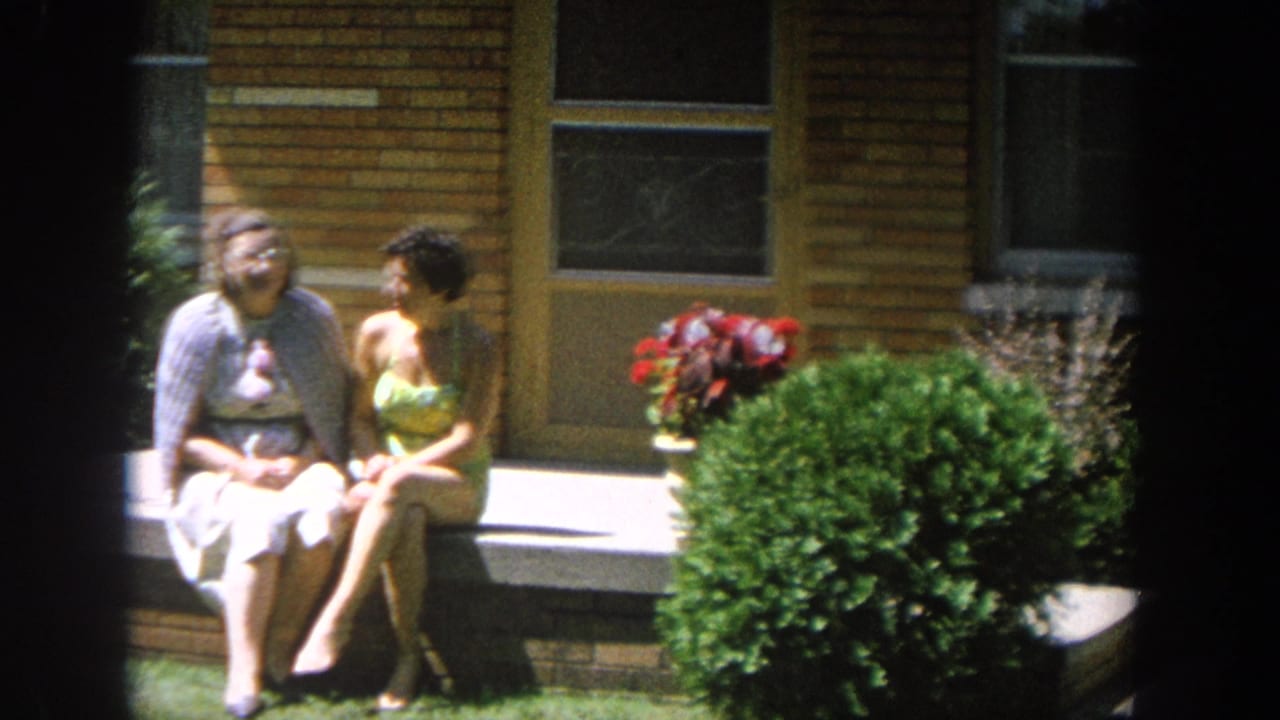 In 1964 Wisconsin home-movie footage, two women sit together outside a brick house near shrubs and a red flowering plant.