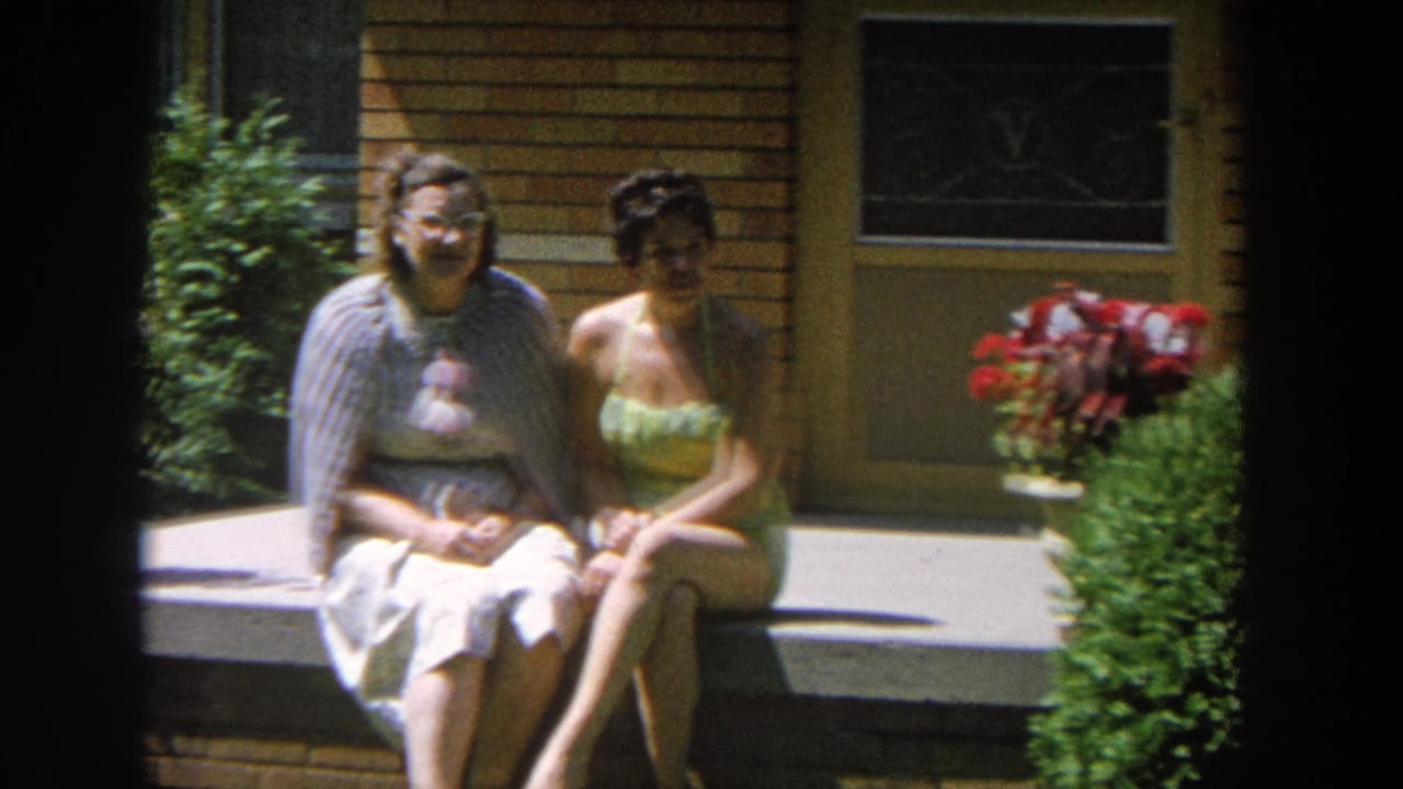 Two women face the camera while seated beside a potted red flowering plant.