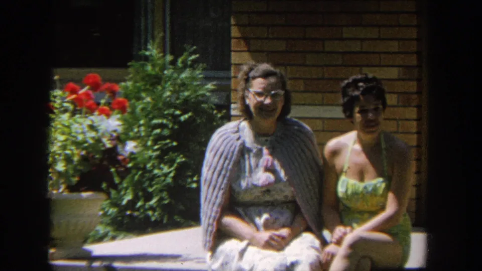 A closer 8mm frame shows two women seated shoulder to shoulder outside a brick house, with red flowers and greenery nearby.