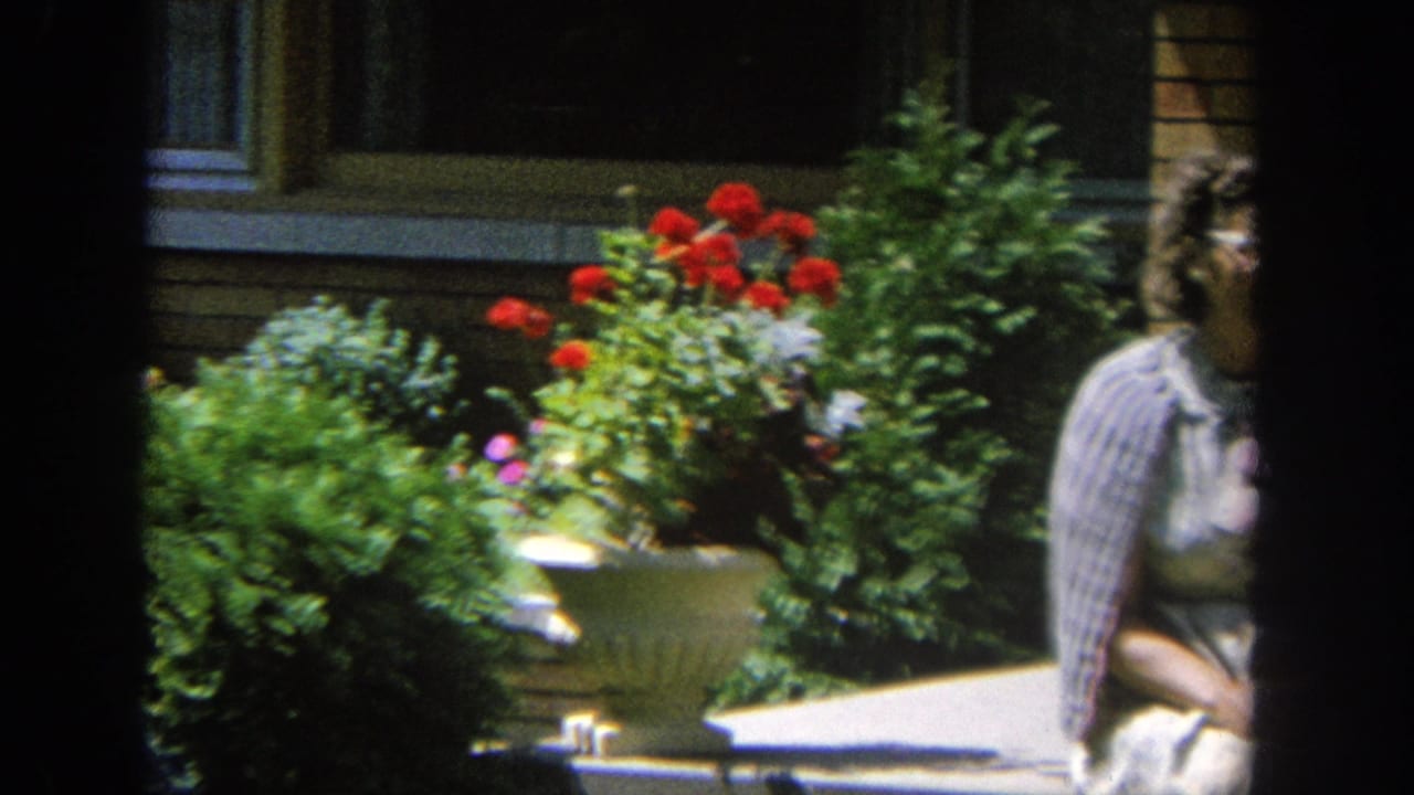 1964 Wisconsin home-movie frame showing a woman in a pale dress near red geraniums and shrubs outside a brick house.