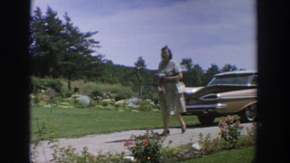1964 Wisconsin yard scene with a woman in a light dress standing near a classic 1960s American car on a gravel driveway.