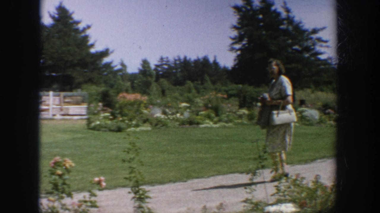 Woman stands on a path near a lawn, garden beds, white fence, and trees.
