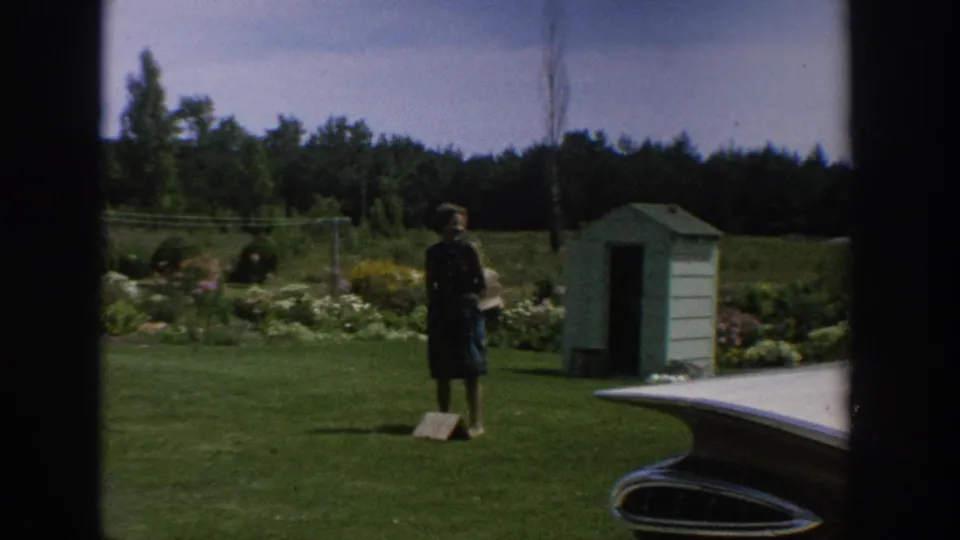 A woman stands in a garden lawn area near a small shed, flower beds, and the rear of a vintage car in the 1964 Wisconsin home-movie scene.