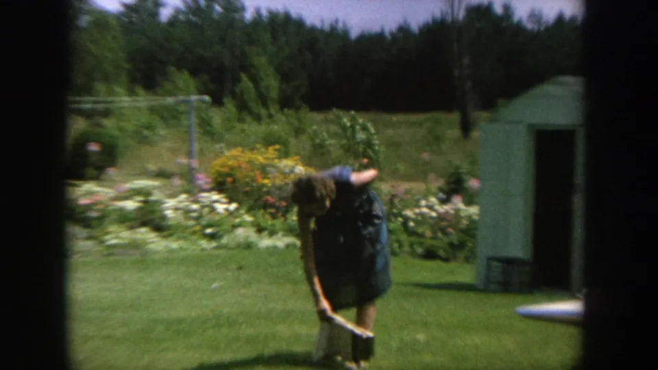 The woman bends toward an object on the lawn near garden flowers, a cement path, and a small green shed in the Wisconsin home-movie footage.